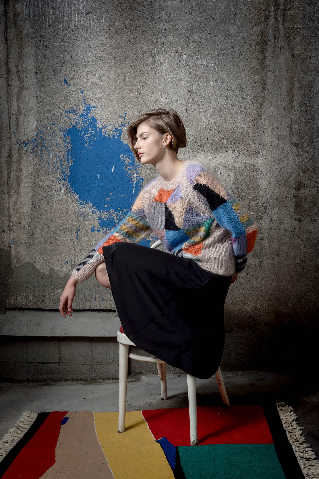 Woman wearing a mohair sweater sitting on chair. The sweater is beige and is covered with colourful abstract colour block patterns on most of its surface.
