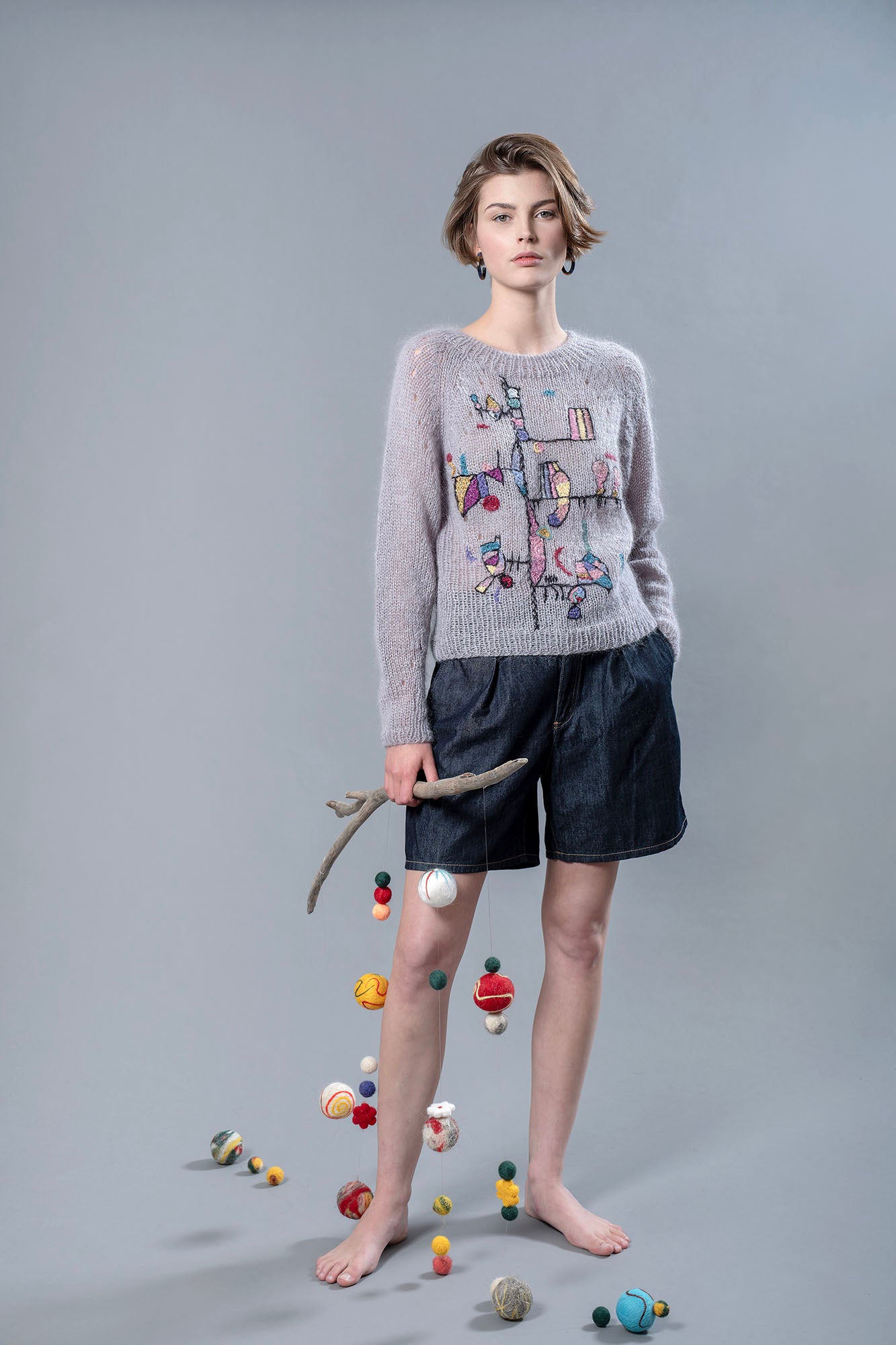 Woman wearing a grey mohair sweater and holding in her right hand a wooden stick from which wool balls are hanging. The sweater has colourful abstract embroidered patterns and shapes on the front..
