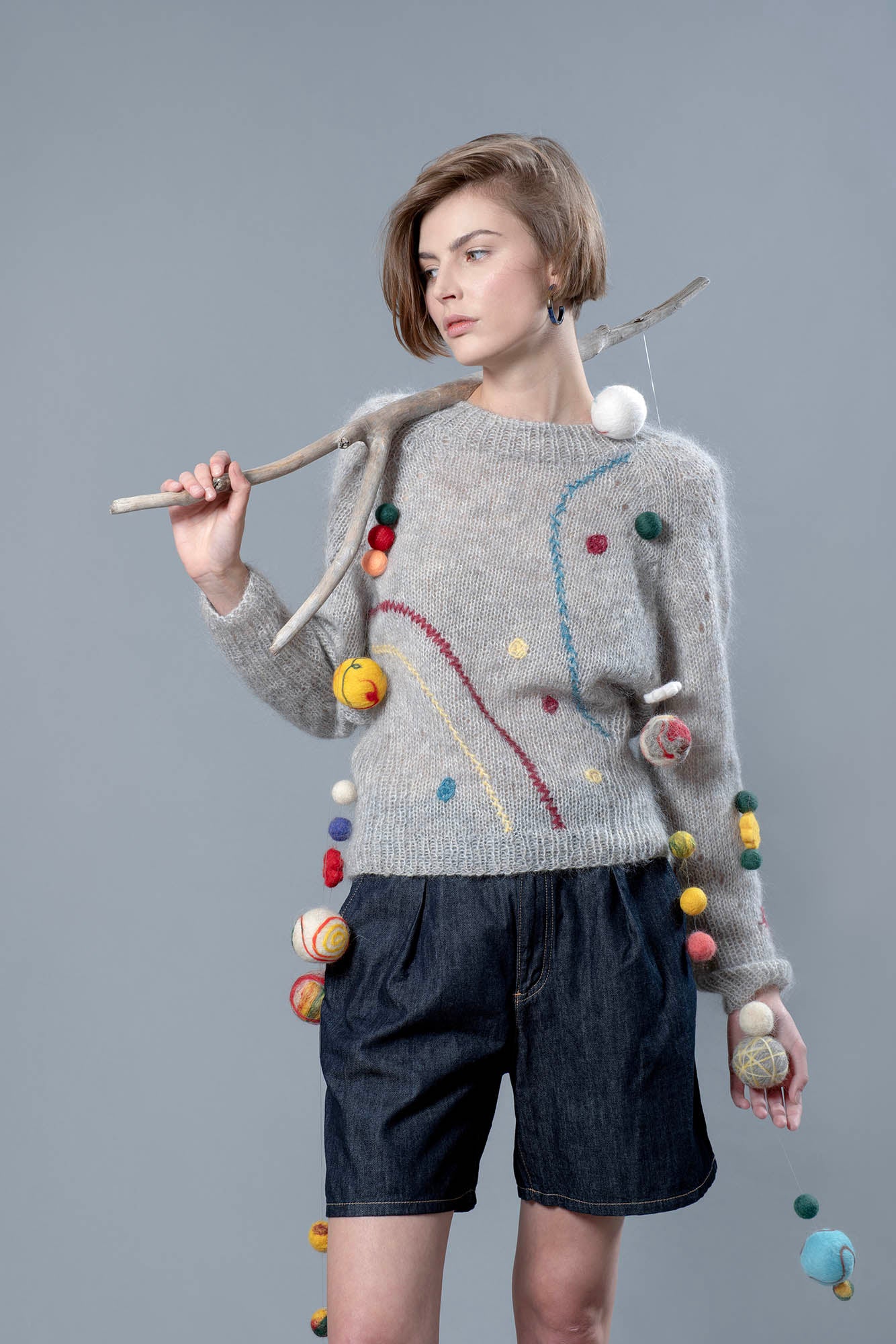 Woman wearing grey mohair sweater and holding over her shoulder a wooden stick from which wool balls are hanging. The sweater has 3 colourful embroidered line running along it and a few embroidered dots.