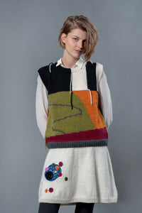 Thumbnail for Woman wearing white wool knee length skirt and wool vest. The skirt has colourful bubble-like embroideries on the right leg. 