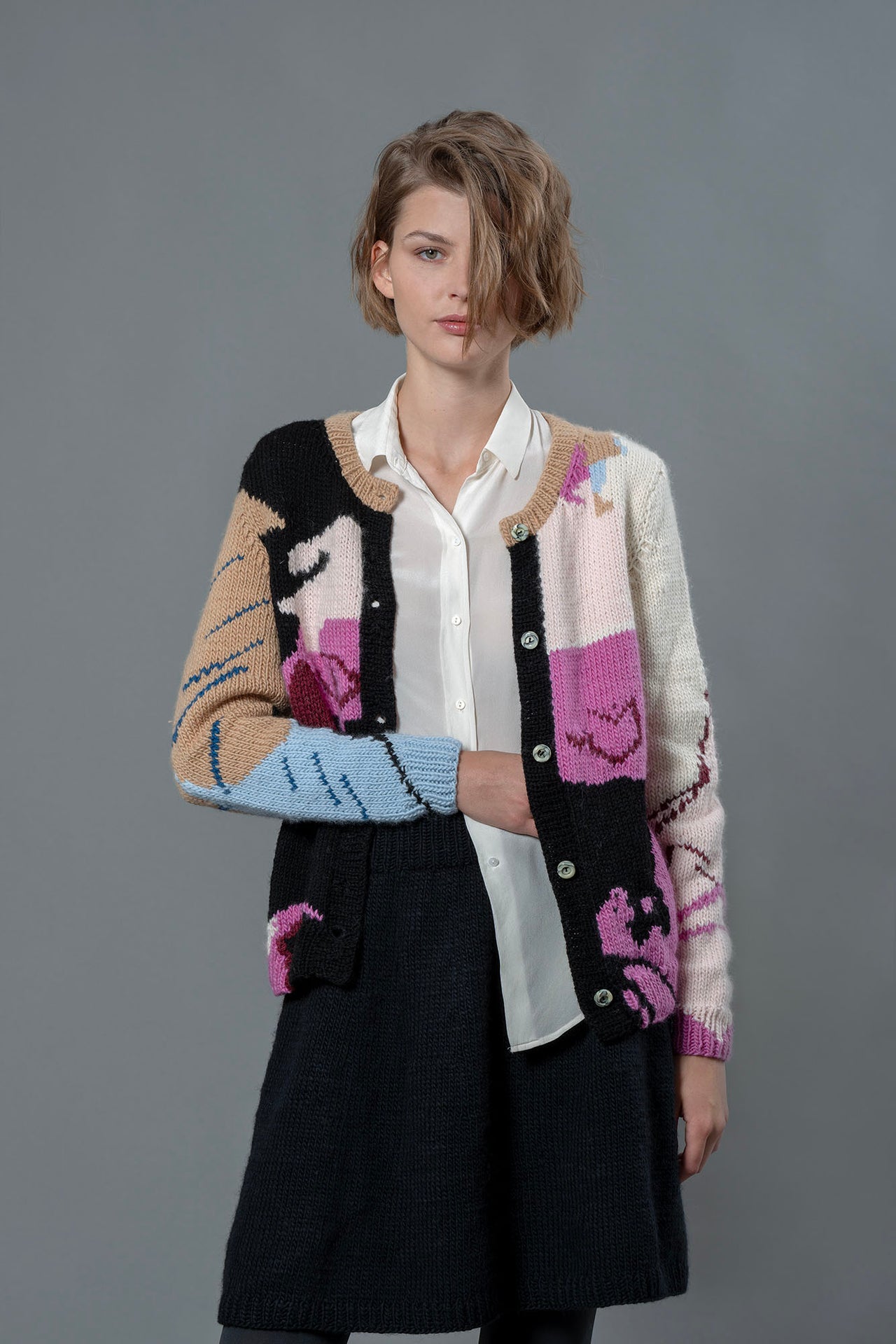 Woman model posing with a colourful wool cardigan. Spots and shapes of fuschia, pastel pink, white black, beige, baby blue and black are on the cardigan. 