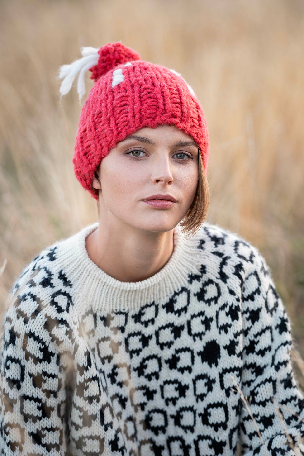 Closeup of a woman sitting in a wheat field. Wearing a red beanie (our Freya beanie) with white embroideries and a wool sweater with a herd of white sheep and a single black sheep on it 