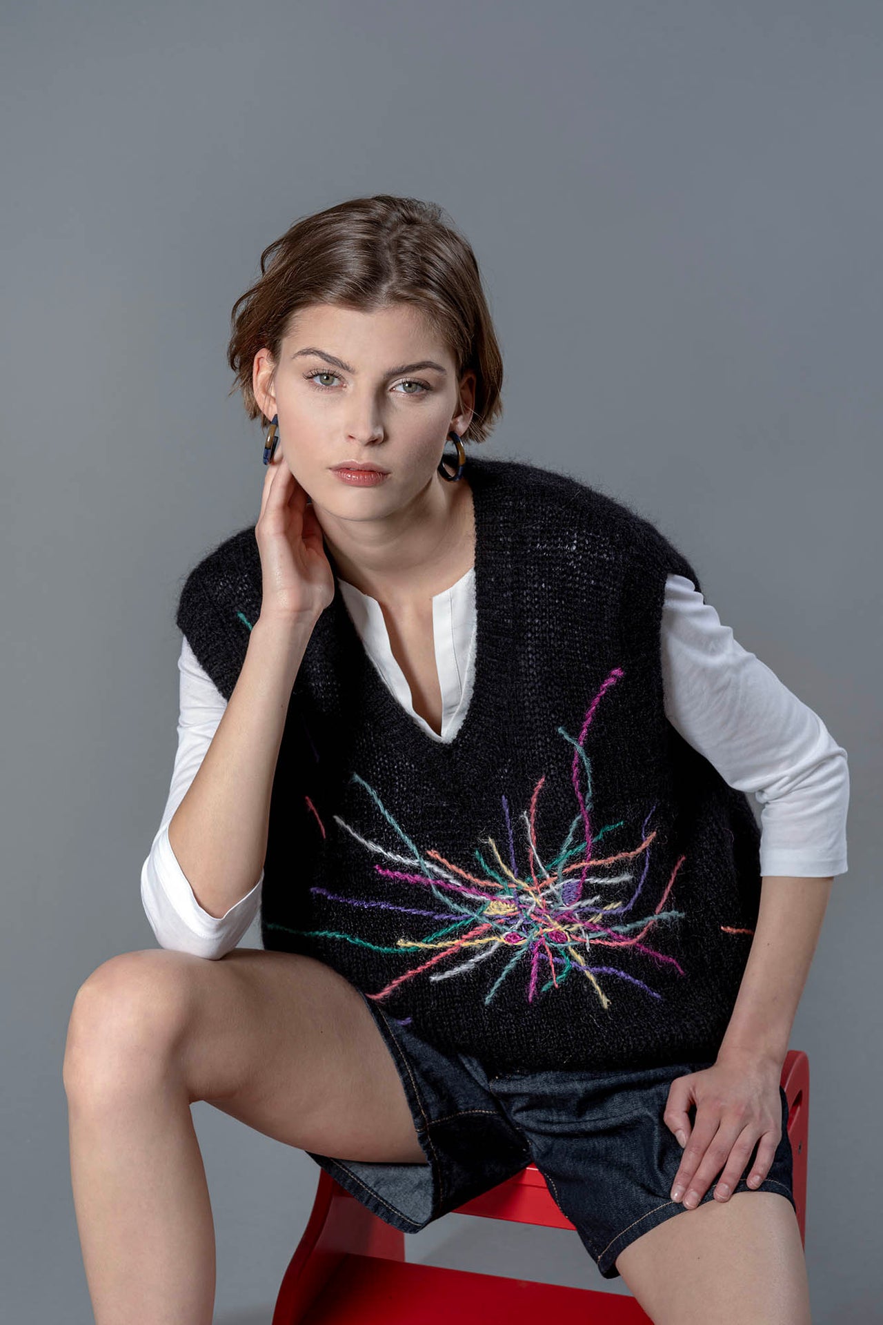 Woman wearing a black mohair vest sitting on a stool. The black vest has colourful embroidered lines going in all directions from the center