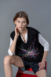 Thumbnail for Woman wearing a black mohair vest sitting on a stool. The black vest has colourful embroidered lines going in all directions from the center