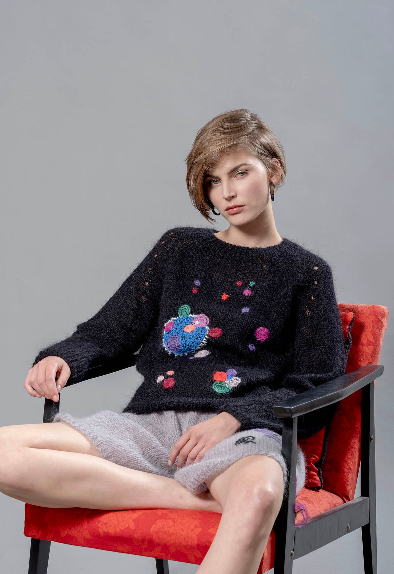 Woman sitting on red chair wearing black mohair sweater and grey mohair shorts. The sweater has colourful abstract star-like embroideries adorning it. 