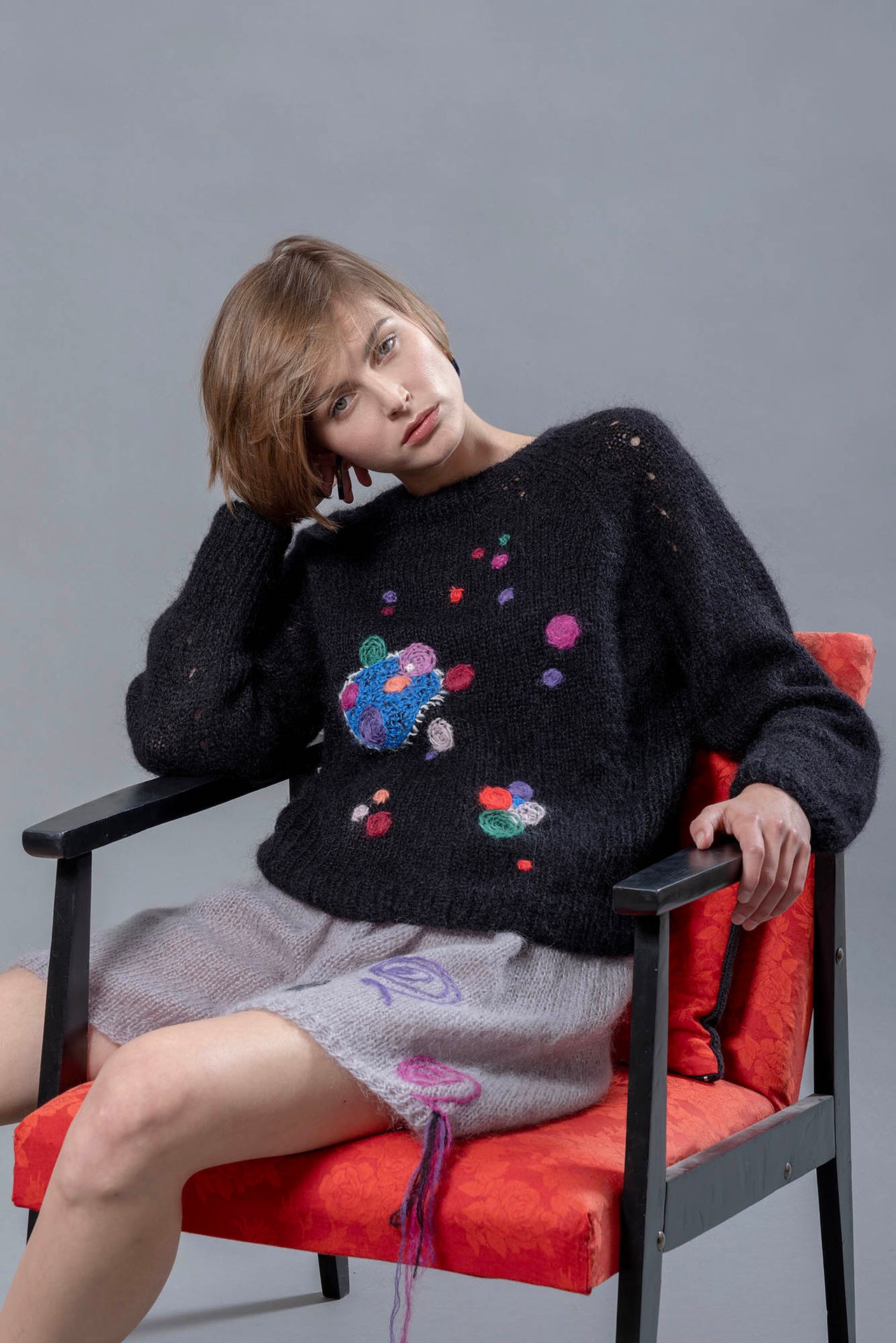 Woman sitting on red chair wearing black mohair sweater and grey mohair shorts. The sweater has colourful abstract star-like embroideries adorning it. 