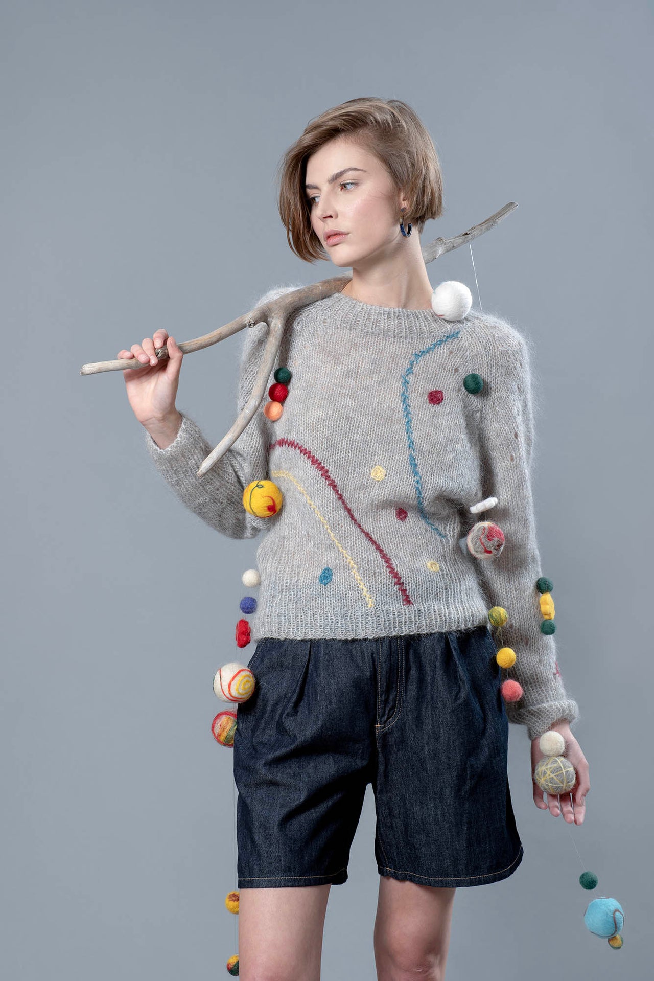 Woman wearing grey mohair sweater and holding over her shoulder a wooden stick from which wool balls are hanging. The sweater has 3 colourful embroidered line running along it and a few embroidered dots. 