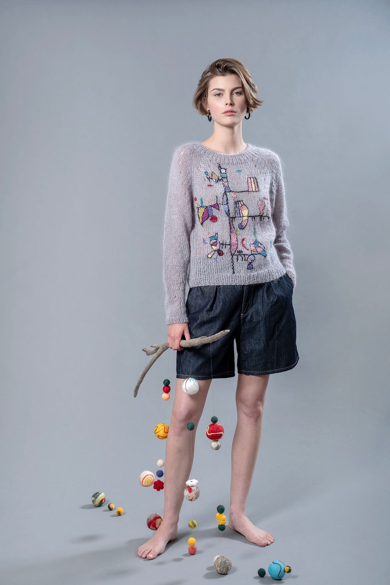 Woman wearing a grey mohair sweater and holding in her right hand a wooden stick from which wool balls are hanging. The sweater has colourful abstract embroidered patterns and shapes on the front.. 