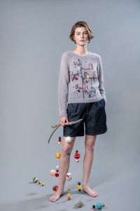 Thumbnail for Woman wearing a grey mohair sweater and holding in her right hand a wooden stick from which wool balls are hanging. The sweater has colourful abstract embroidered patterns and shapes on the front.. 