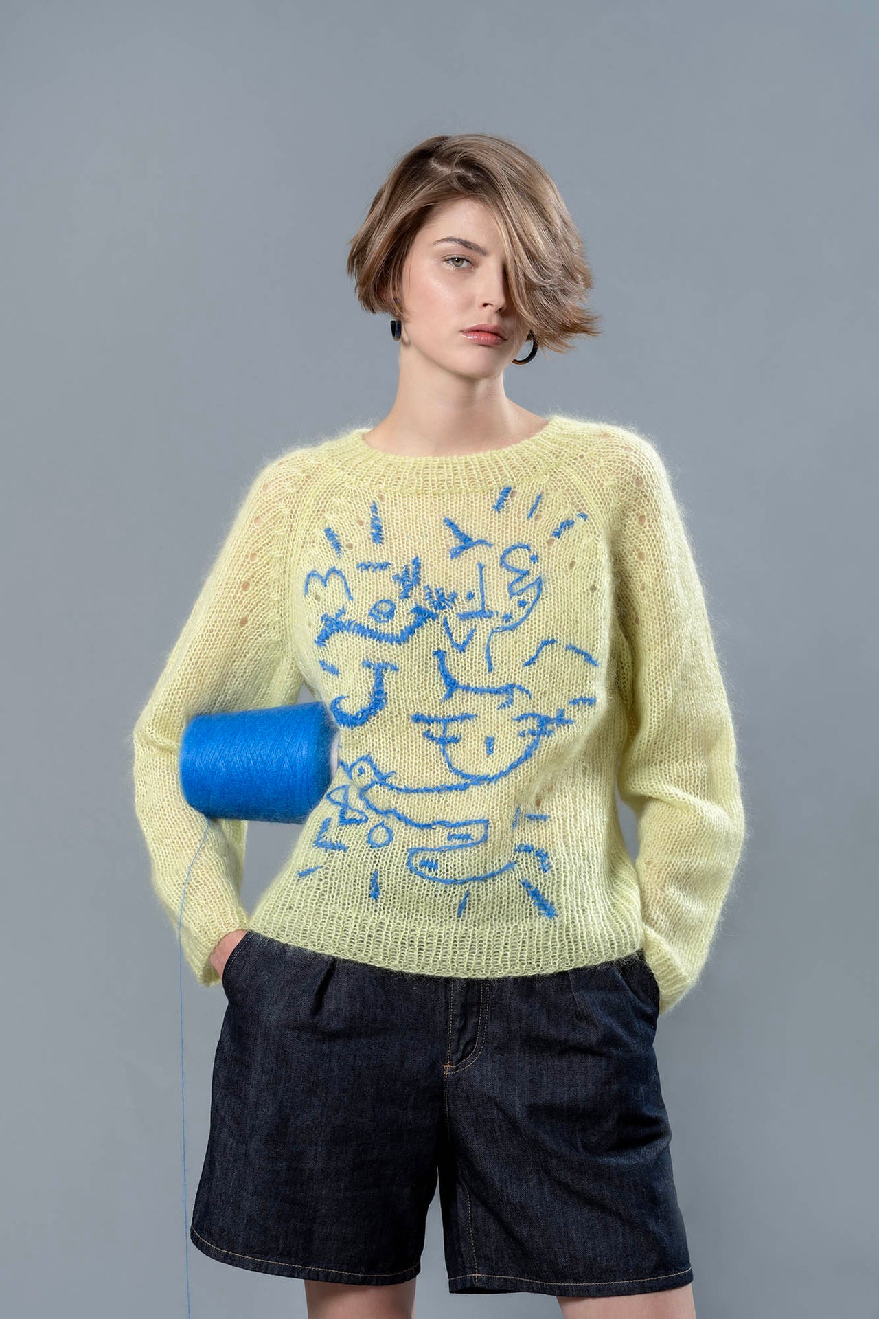 Woman wearing lime coloured mohair sweater and carrying blue wool cone under the arm. The sweater is adorned with sky blue abstract embroidered shapes. 