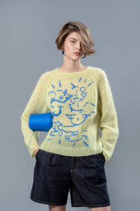 Thumbnail for Woman wearing lime coloured mohair sweater and carrying blue wool cone under the arm. The sweater is adorned with sky blue abstract embroidered shapes. 