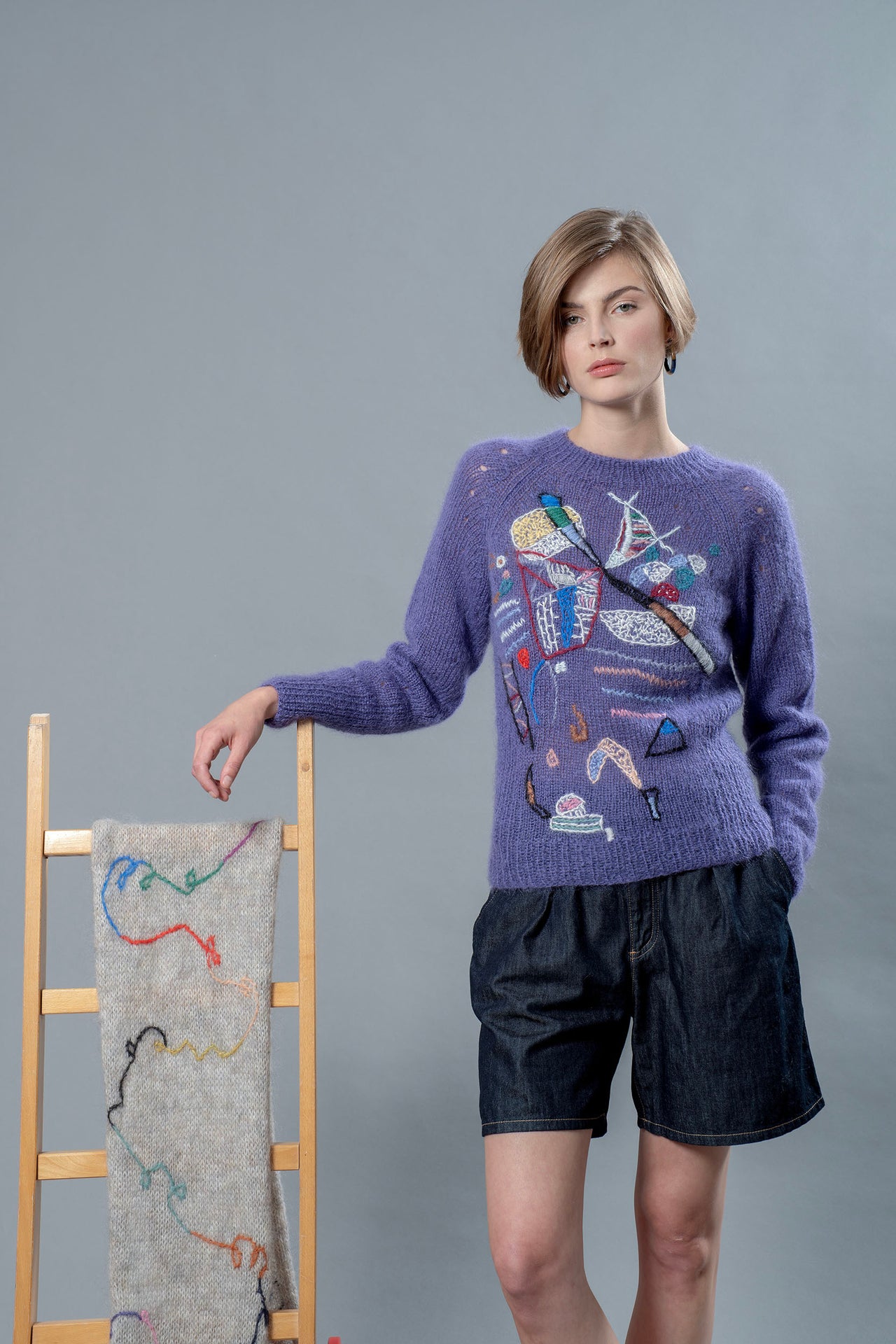 Woman wearing a purple mohair sweater leaning on a chair on which a grey scarf is hanging. The sweater has colourful abstract motifs embroidered over it. 