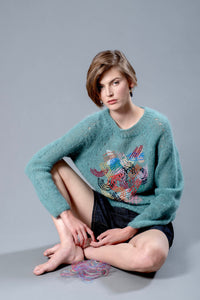 Thumbnail for Woman sitting on the floor wearing a teal mohair sweater. The sweater has colourful abstract embroidered lines in its center. 