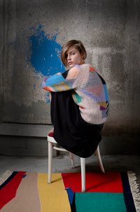 Thumbnail for Woman wearing a mohair sweater sitting on chair. The sweater is beige and is covered with colourful abstract colour block patterns on most of its surface.