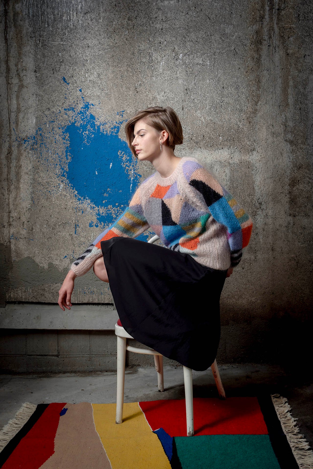 Woman wearing a mohair sweater sitting on chair. The sweater is beige and is covered with colourful abstract colour block patterns on most of its surface.
