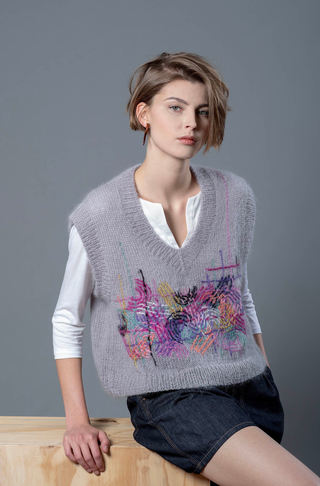 Woman sitting on wood box wearing grey mohair vest. The vest is adorned with colourful embroidered abstract motifs in its center. 