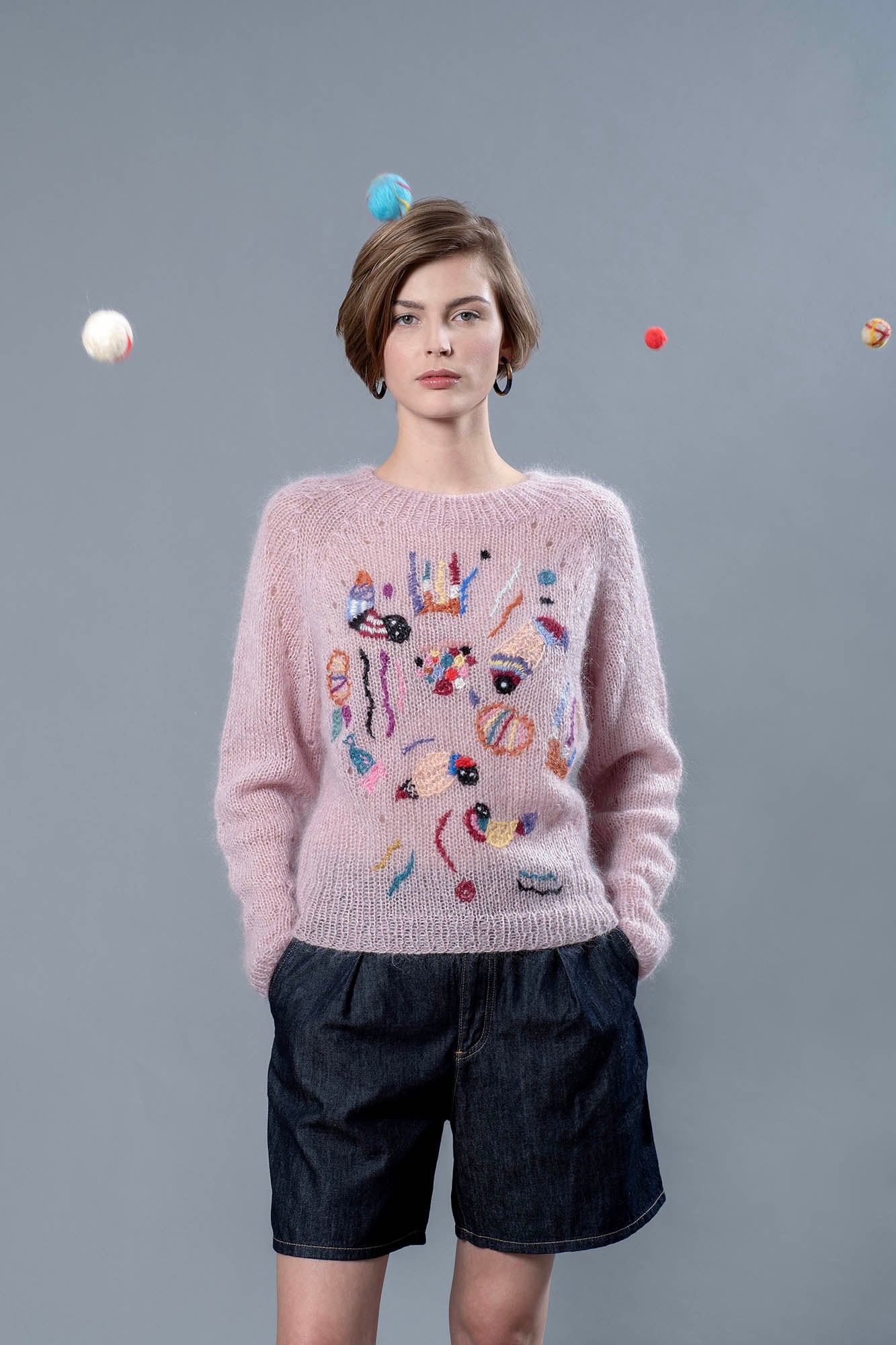 Woman wearing pink mohair sweater with wool balls floating around her head. The mohair sweater is light pink with some colourful abstract embroideries adorning it. 