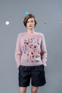 Thumbnail for Woman wearing pink mohair sweater with wool balls floating around her head. The mohair sweater is light pink with some colourful abstract embroideries adorning it. 