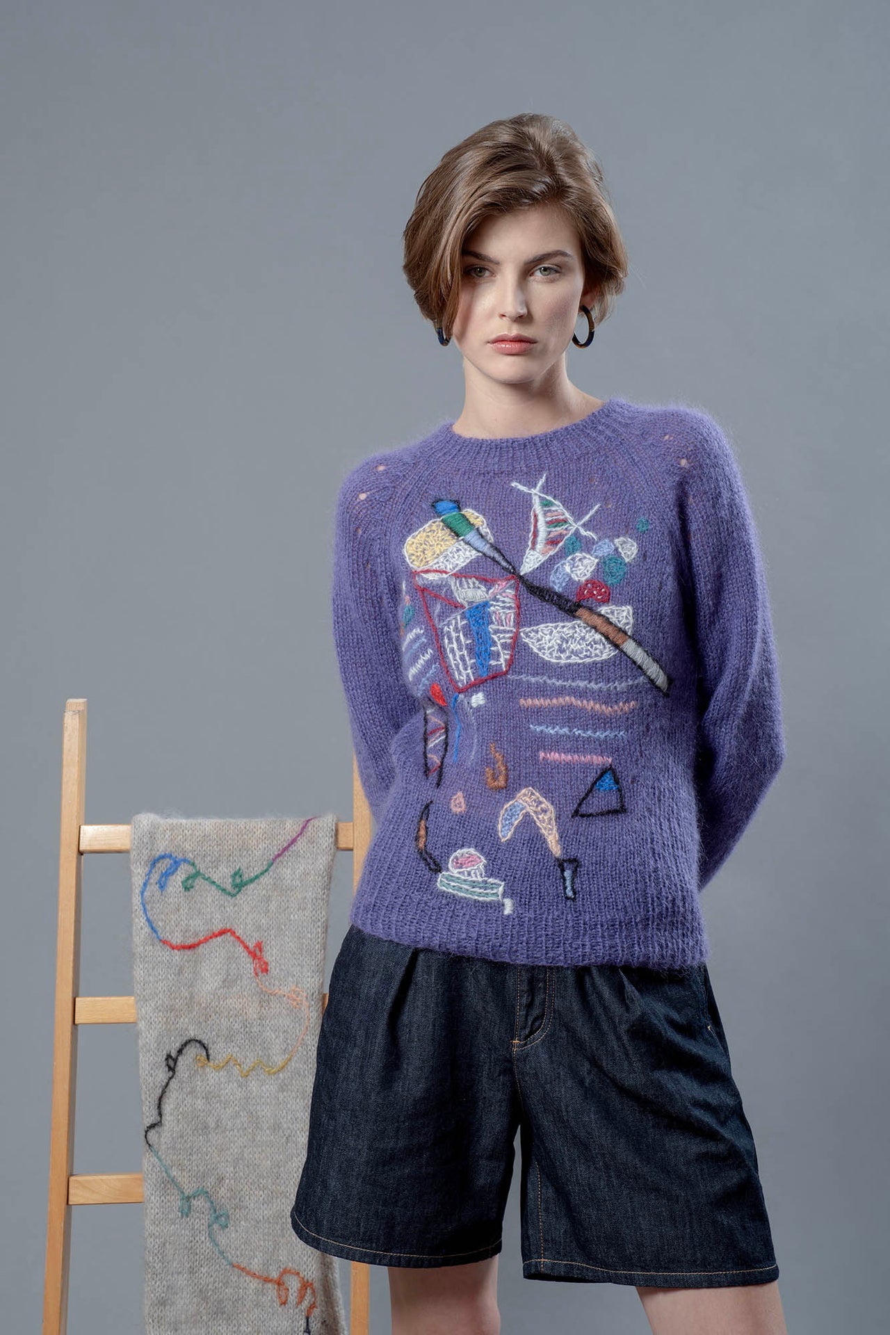 Woman wearing a purple mohair sweater leaning on a chair on which a grey scarf is hanging. The sweater has colourful abstract motifs embroidered over it. 
