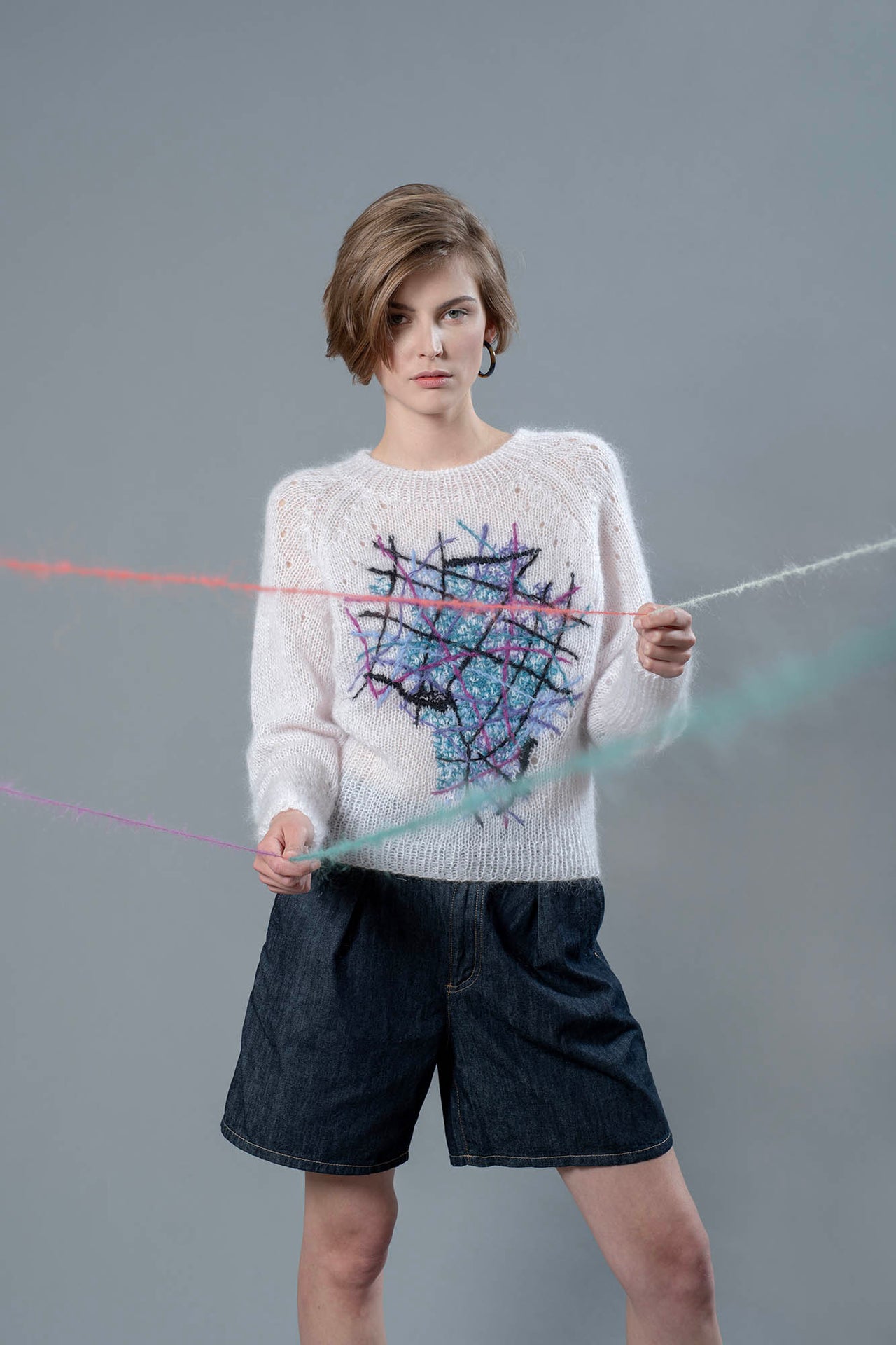 Woman wearing light pink mohair sweater and holding wool yarns going towards camera. The sweater has colourful embroidered lines forming an abstract shape in the center. 