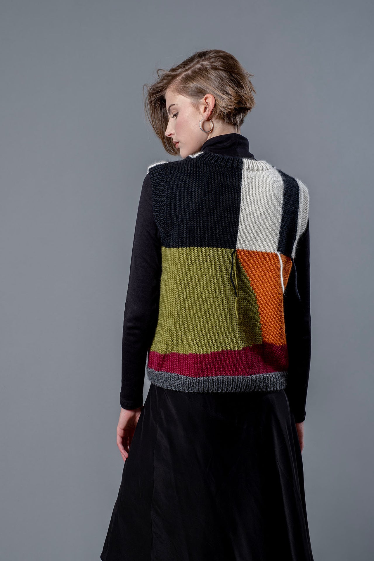 Women wearing wool vest from behind. Wool vest with green, orange, black, white and red colour block design and hanging threads.