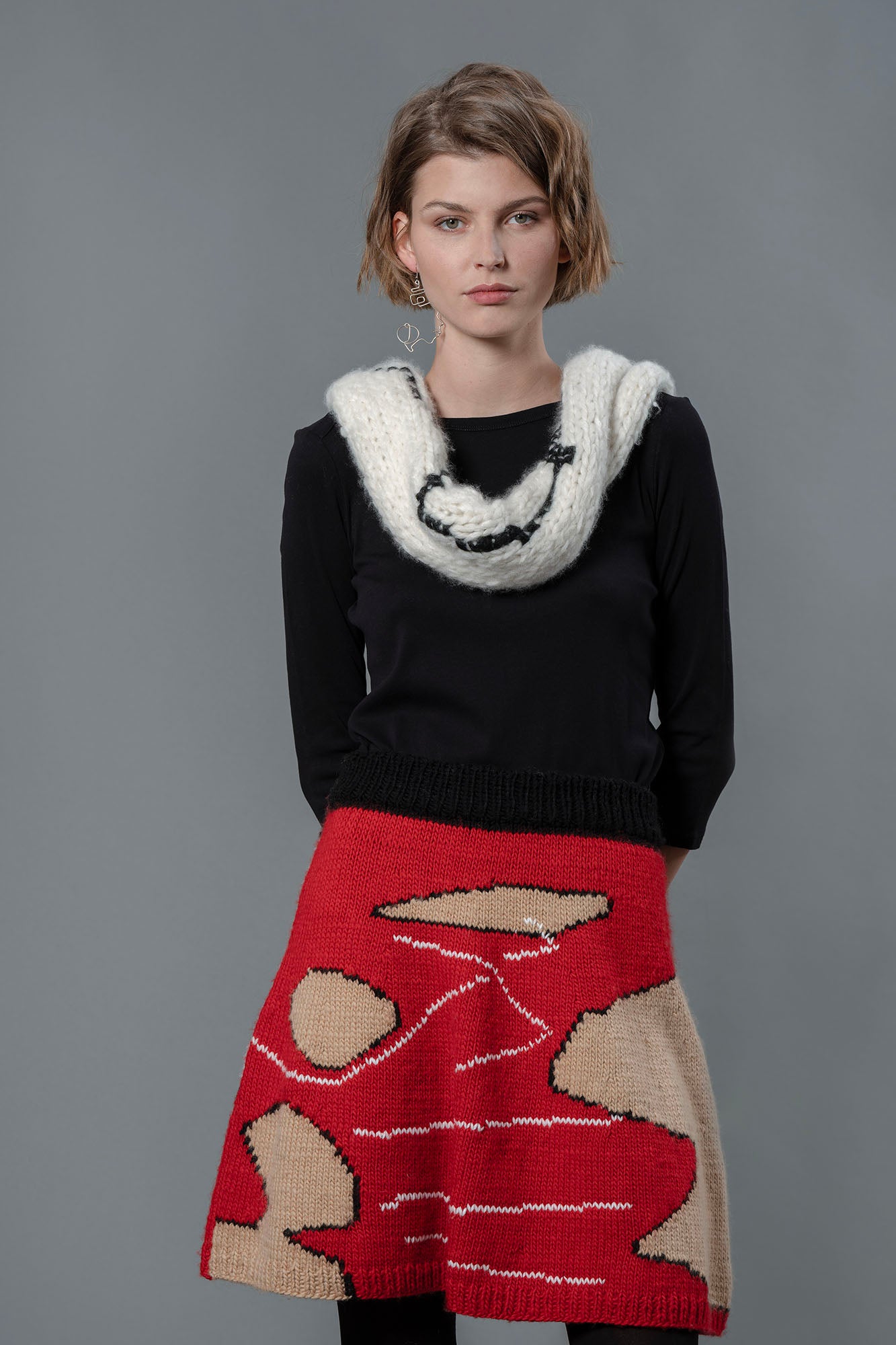 Woman wearing white wool scarf, black top and red wool skirt. The skirt is predominantly red with abstract shapes of beige and white lines. 