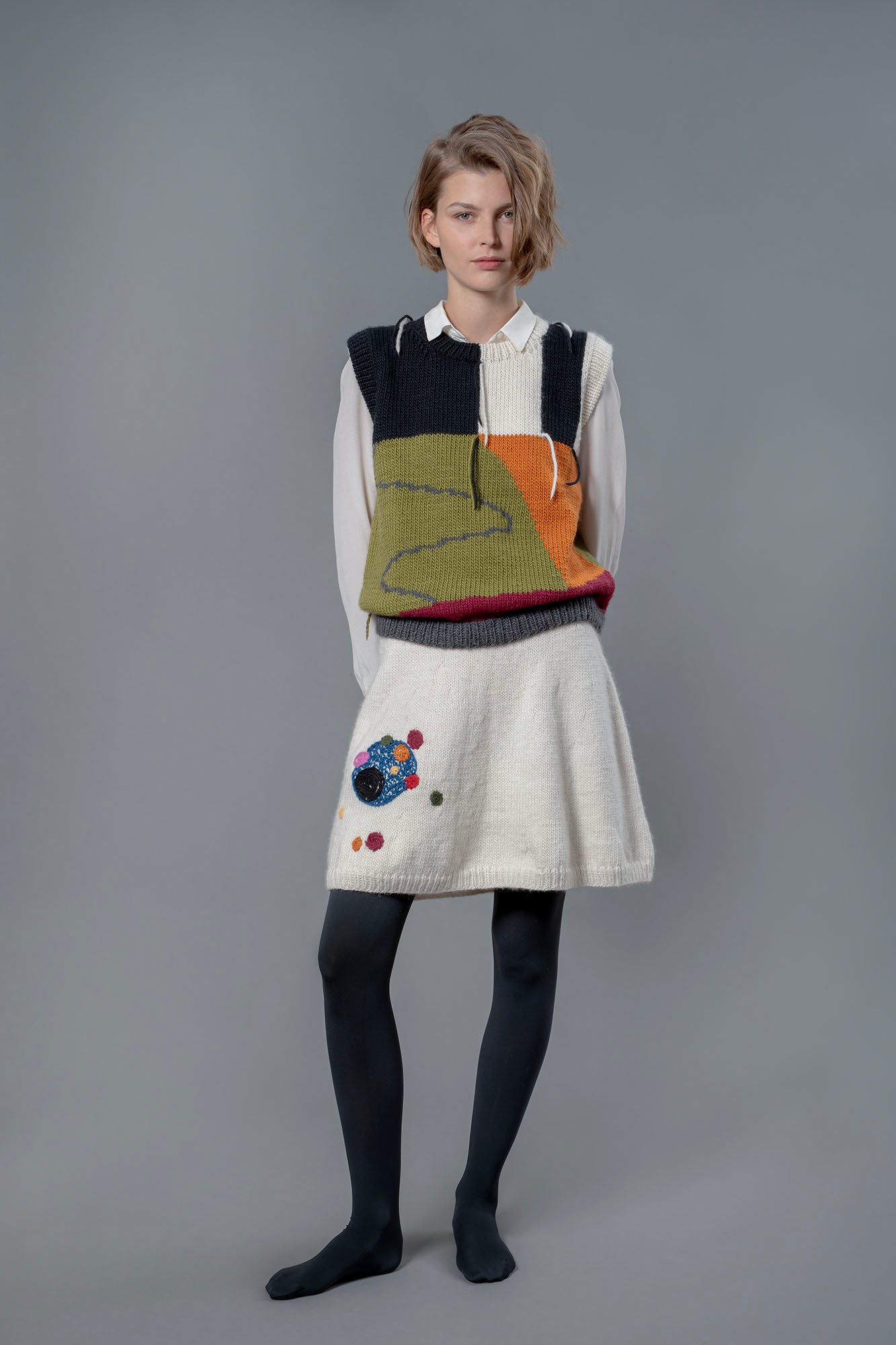 Woman wearing white wool knee length skirt and wool vest. The skirt has colourful bubble-like embroideries on the right leg. 