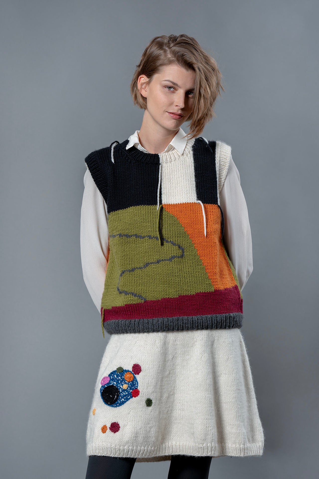 Woman wearing white wool knee length skirt and wool vest. The skirt has colourful bubble-like embroideries on the right leg. 
