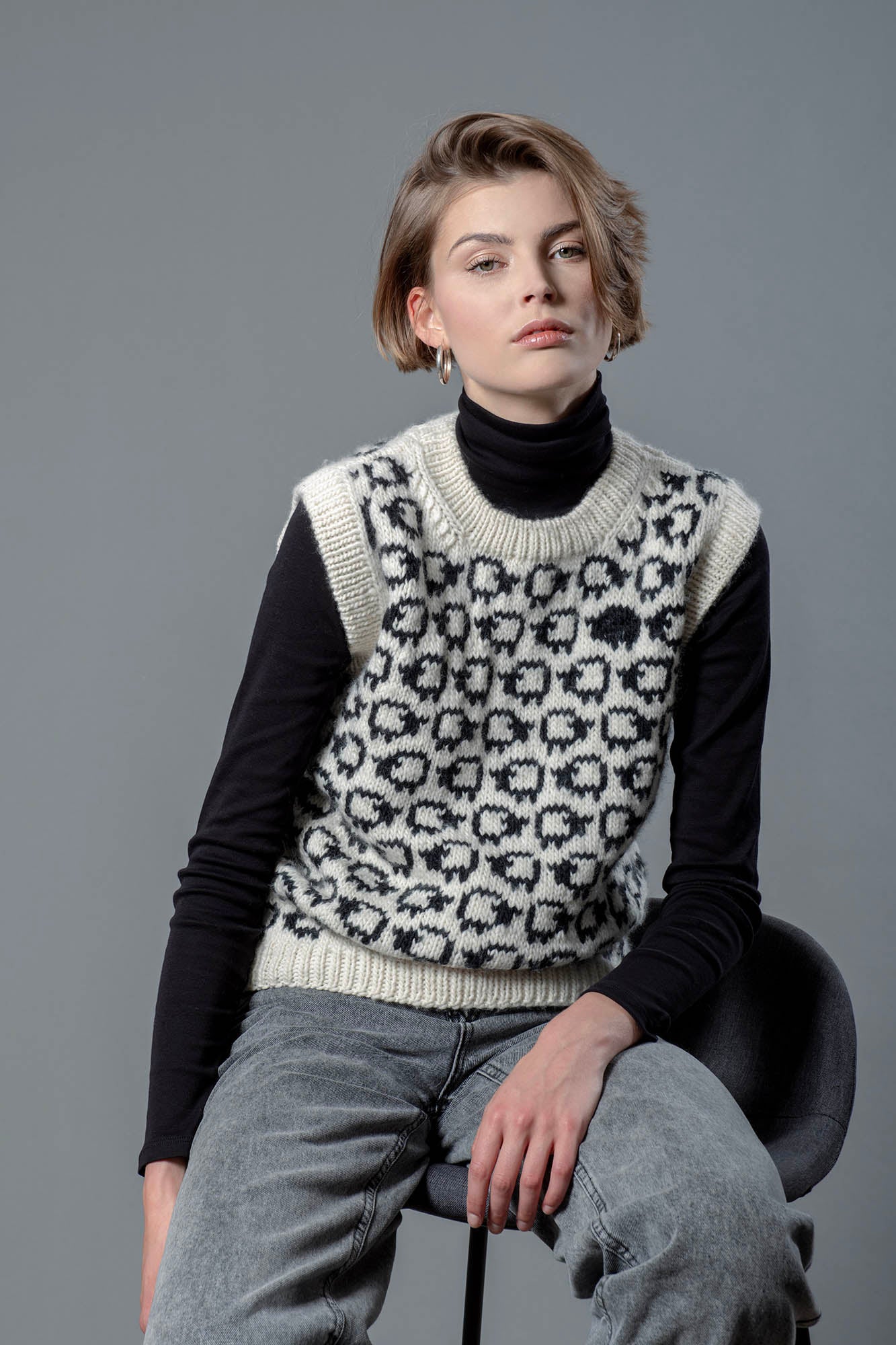 Woman wearing a wool vest. Vest has a herd of white sheep going all around it, there is one single black sheep lost in the herd. 