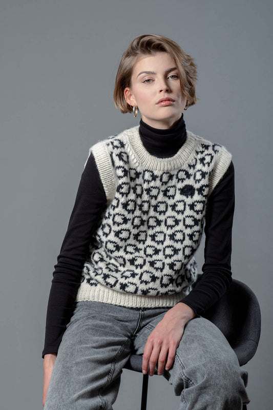 Woman wearing a wool vest. Vest has a herd of white sheep going all around it, there is one single black sheep lost in the herd. 