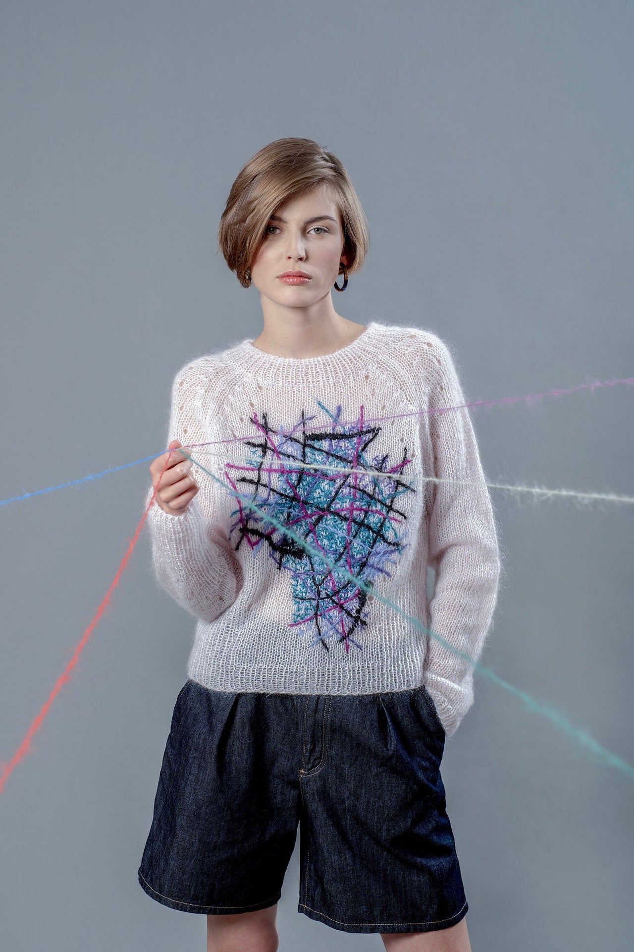 Woman wearing light pink mohair sweater and holding wool yarns going towards camera. The sweater has colourful embroidered lines forming an abstract shape in the center. 